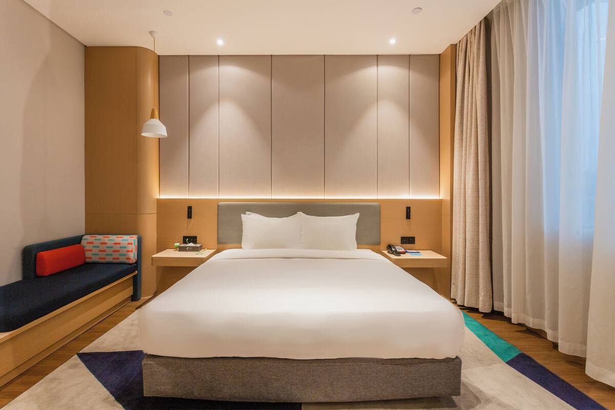 A modern hotel room with a large bed, bedside tables, a nightstand, a sofa with a red cushion, and a large window with curtains.