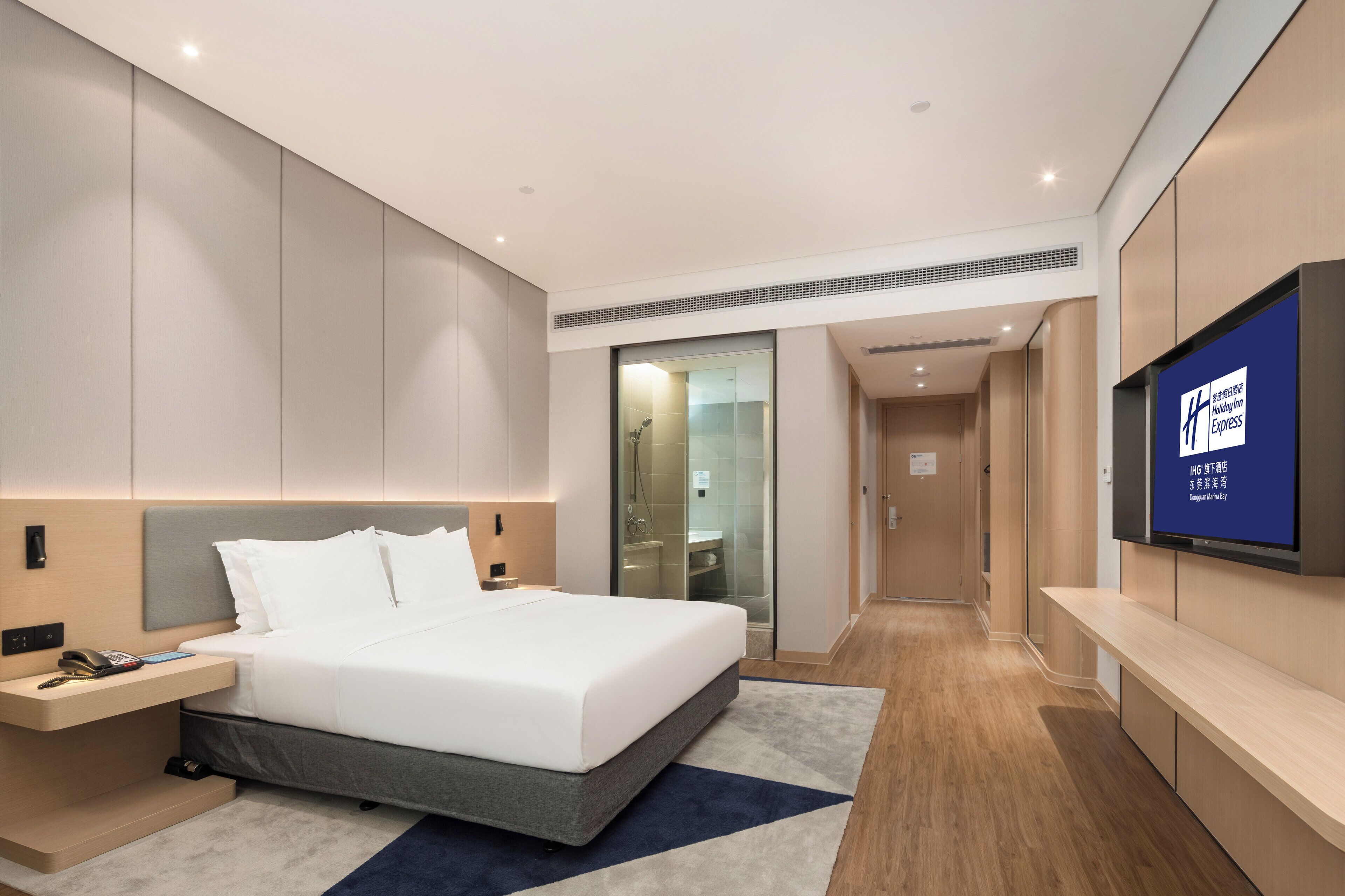 A modern hotel room with a large bed, a bedside table with a phone, a TV mounted on the wall, and a bathroom visible through a glass door.