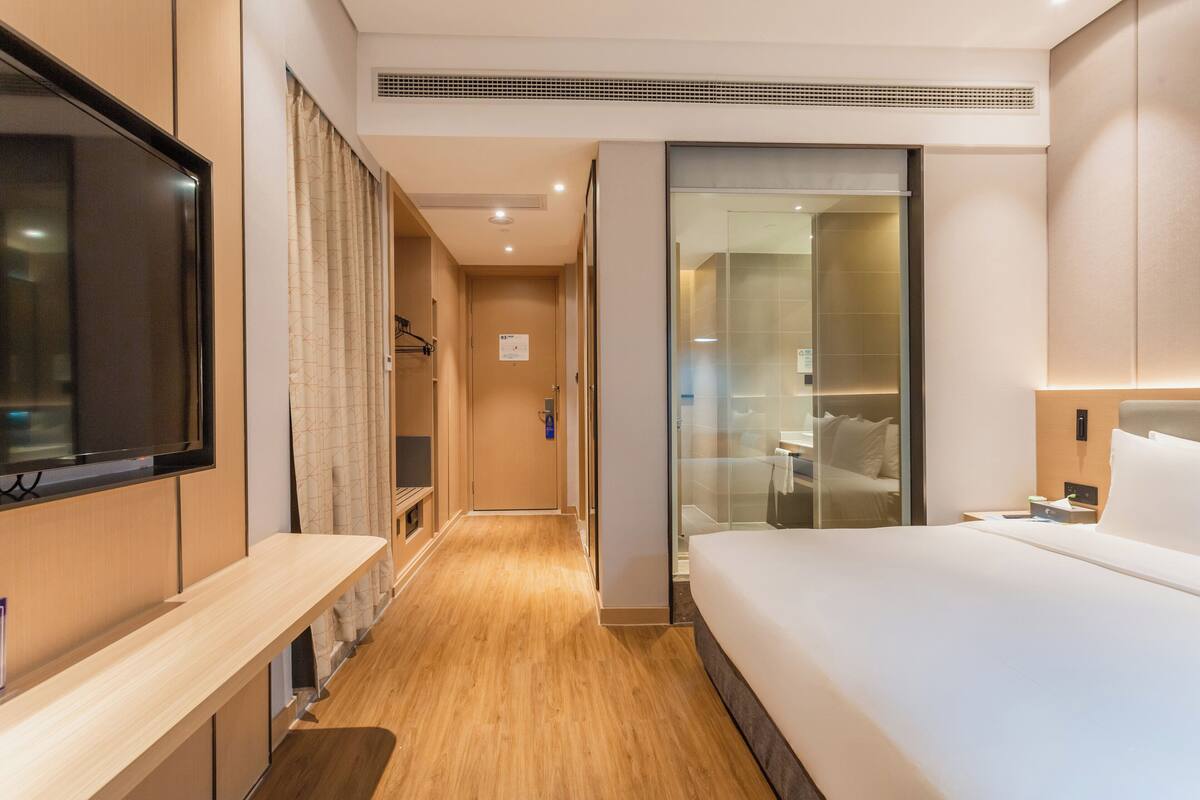 A modern hotel room with a large bed, a TV mounted on the wall, a wooden floor, and a glass shower enclosure.