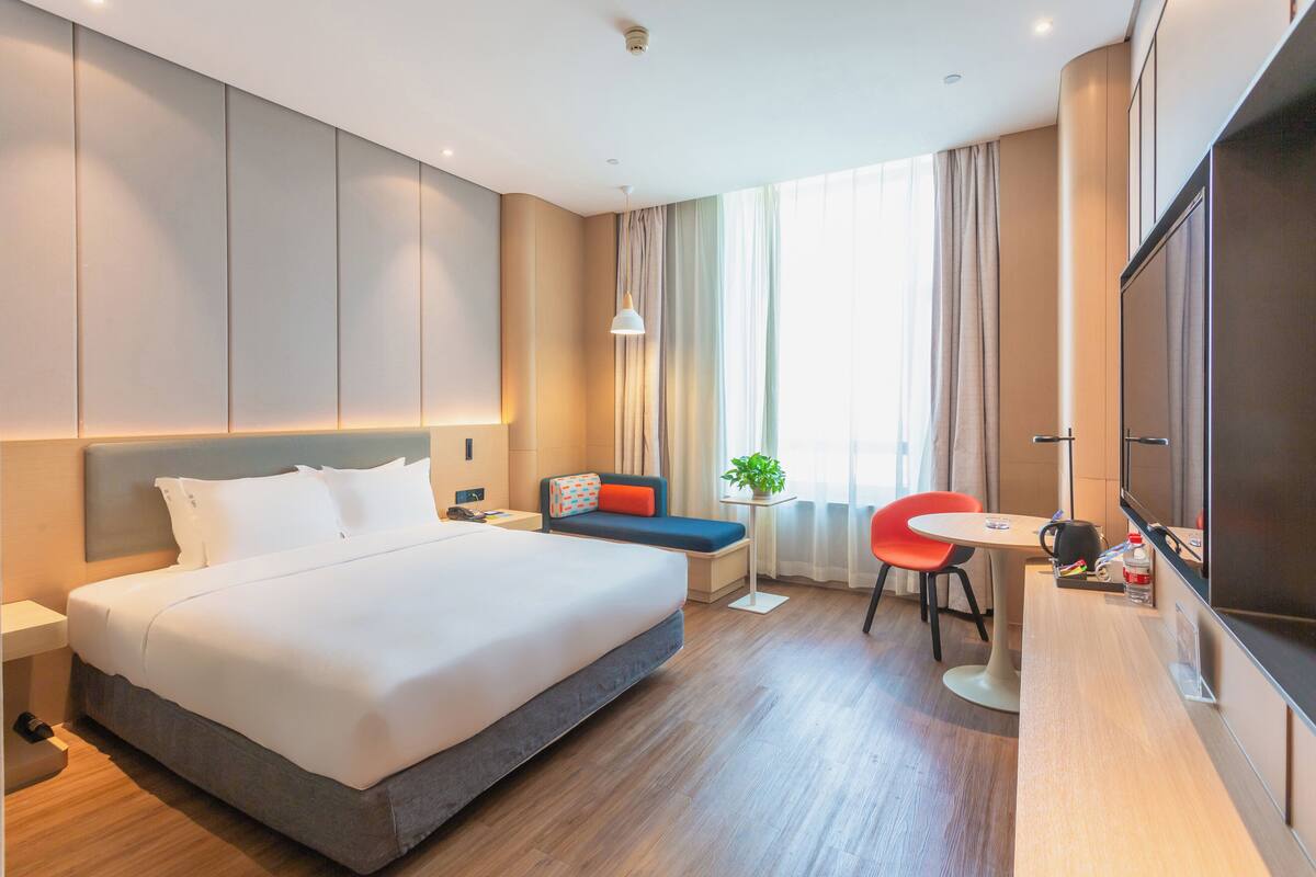 Habitació d'hotel moderna amb un llit gran, una escriptori, una cadira, un televisor i una taula petita amb una planta.