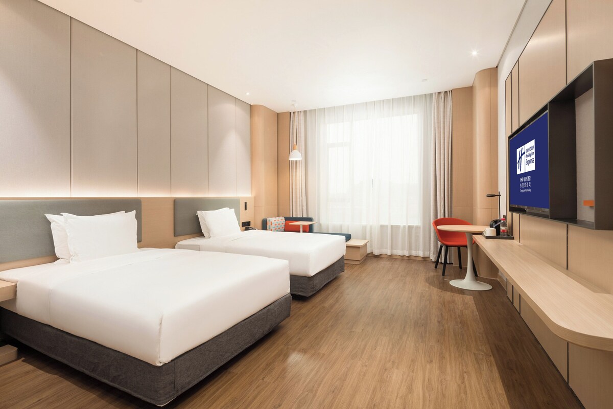 A hotel room with two beds, a desk, a chair, and a TV on the wall.