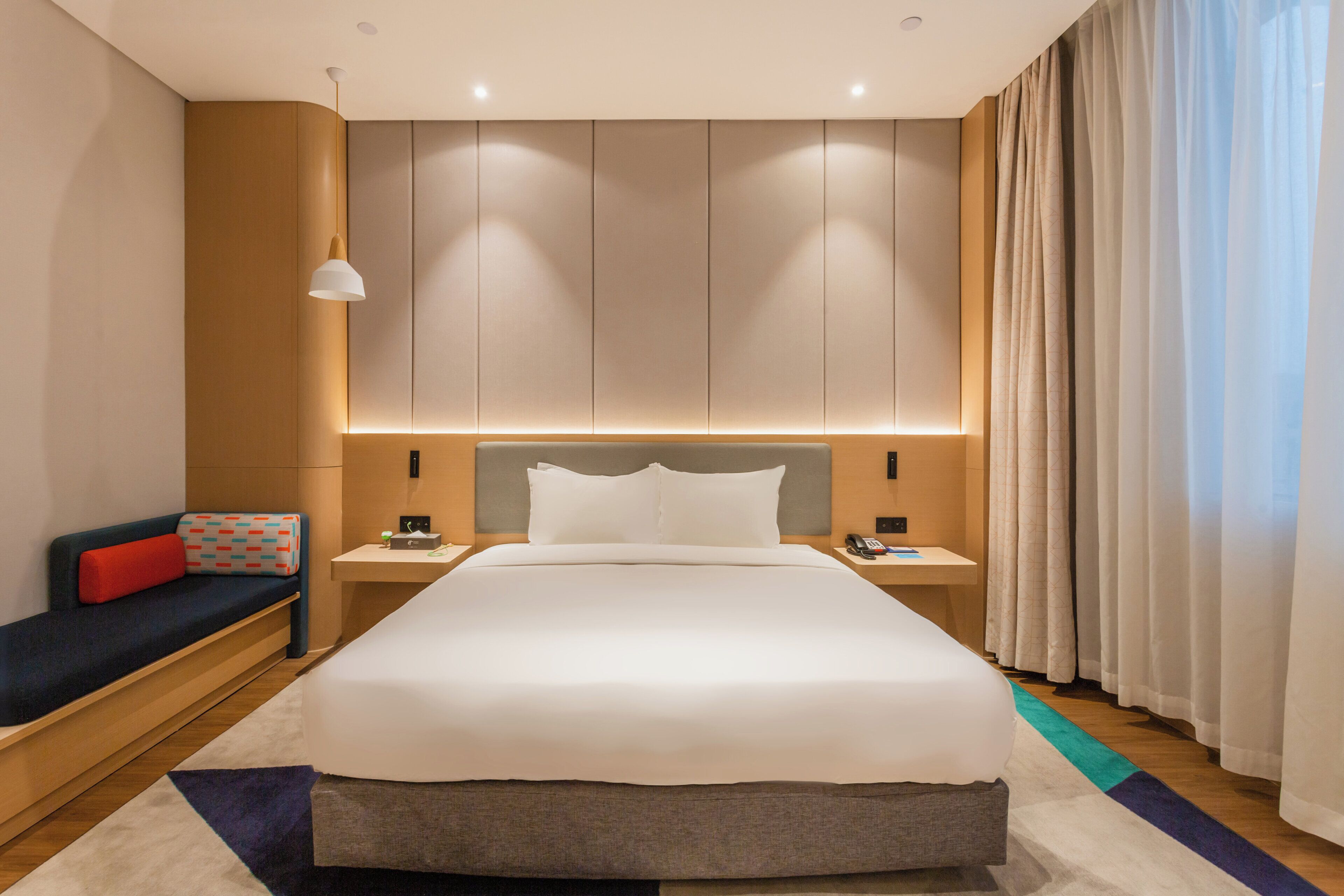 A modern hotel room with a large bed, bedside tables, a nightstand, a sofa with a red cushion, and a large window with curtains.