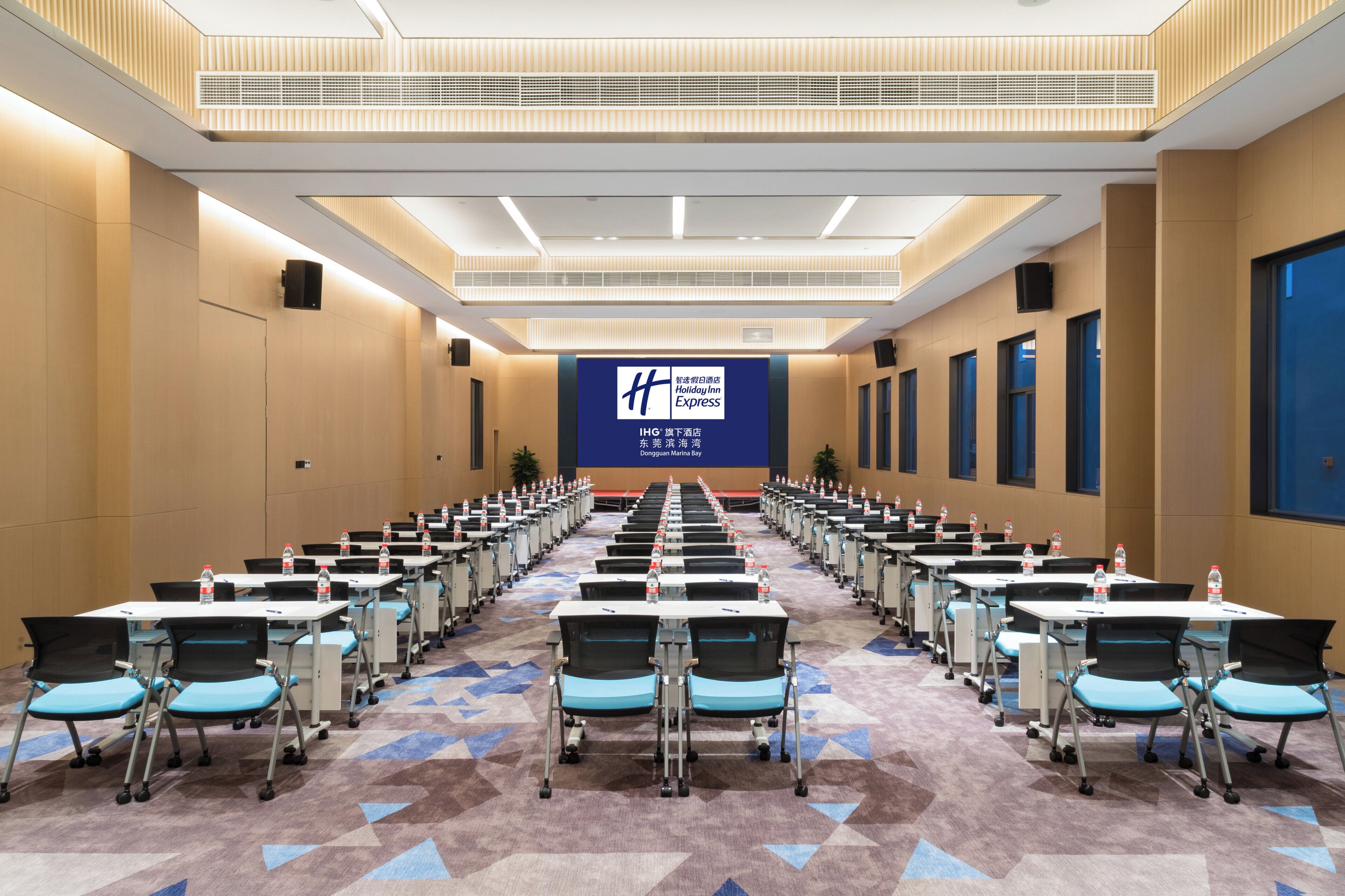 A conference room with rows of tables and chairs, a large screen at the front, and a patterned carpet.