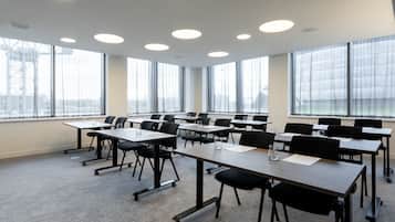 Un salón de conferencias con varias mesas y sillas, ventanas amplias con cortinas e iluminación en el techo.
