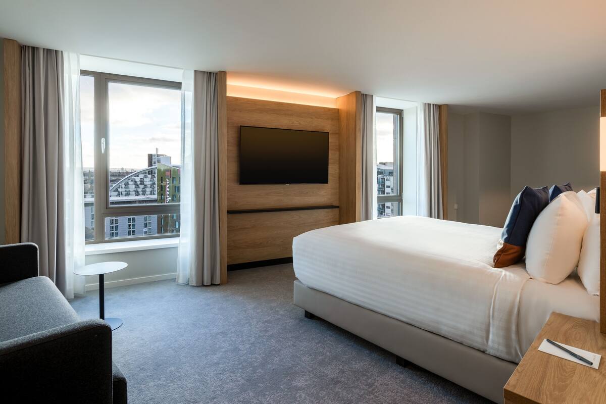 Un hotel moderno con una cama grande, un televisor montado en un panel de madera, una mesita redonda y vistas a la ciudad a través de amplios ventanales.