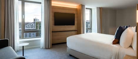 A modern hotel room with a large bed, a TV mounted on a wooden panel, a small round table, and a view of the city through large windows.