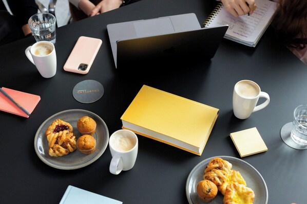 Una mesa con una computadora portátil, un teléfono inteligente, un cuaderno, un libro amarillo, dos platos de pasteles y dos tazas de café.