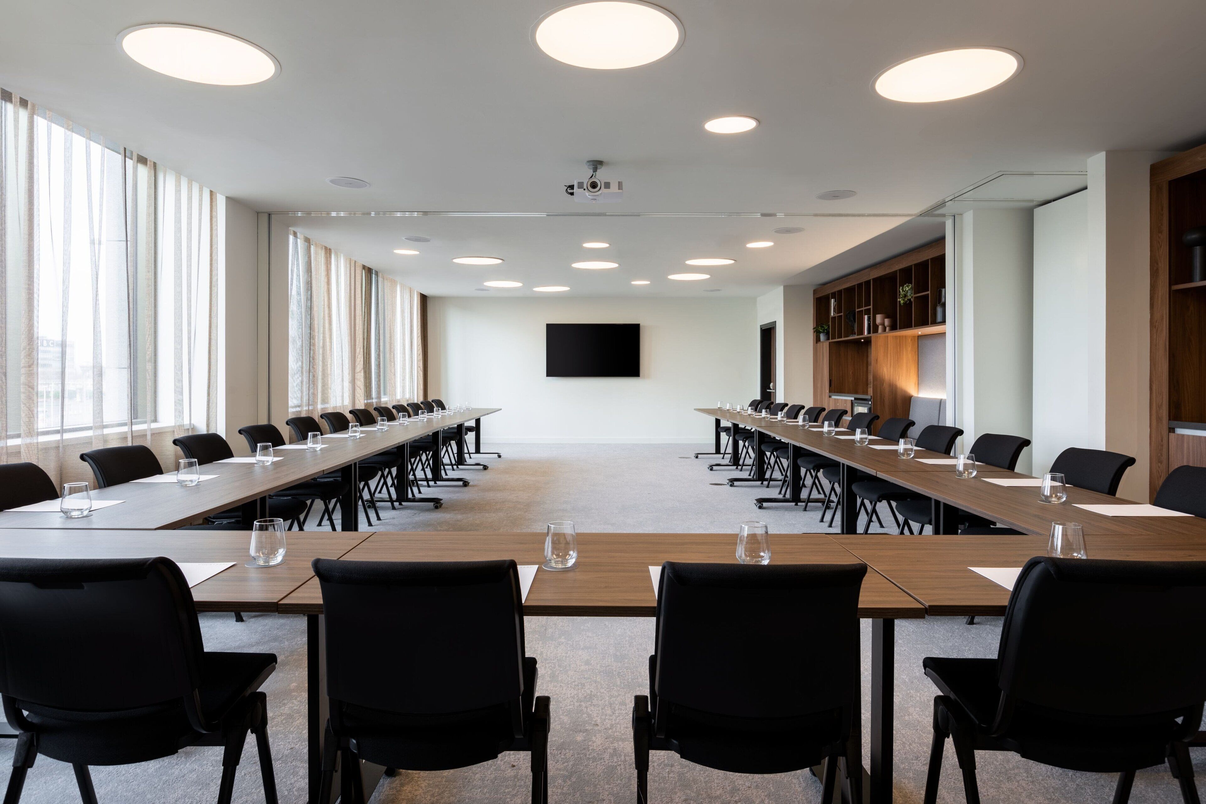A conference room with long tables, black chairs, and a large screen on the wall.
