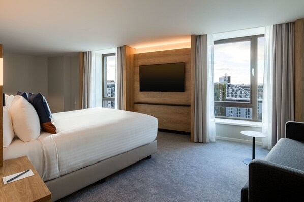Habitación de hotel con una cama grande, un televisor de pantalla plana montado en la pared, una mesita redonda y vistas a la ciudad a través de amplios ventanales.