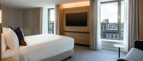 A hotel room with a large bed, a flat-screen TV mounted on the wall, a small table, and a view of the city through the window.