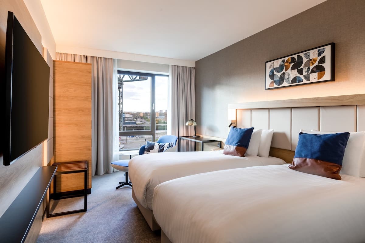 A hotel room with two beds, a desk, a chair, a TV, and a large window with curtains.
