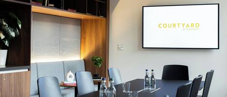 Une salle de conférence dotée d’un grand écran affichant le logo « COURTYARD BY MARRIOTT », d’une longue table entourée de chaises, de bouteilles d’eau et d’une plante en pot.