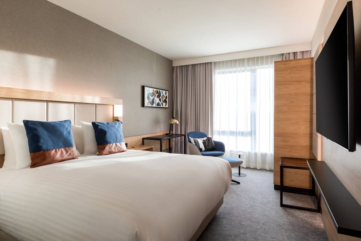 A hotel room with a large bed, a desk, a chair, a TV, and a window with curtains.
