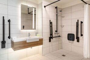 Un baño moderno con tocador de madera, un espejo grande y una ducha a ras de suelo con barras de apoyo.
