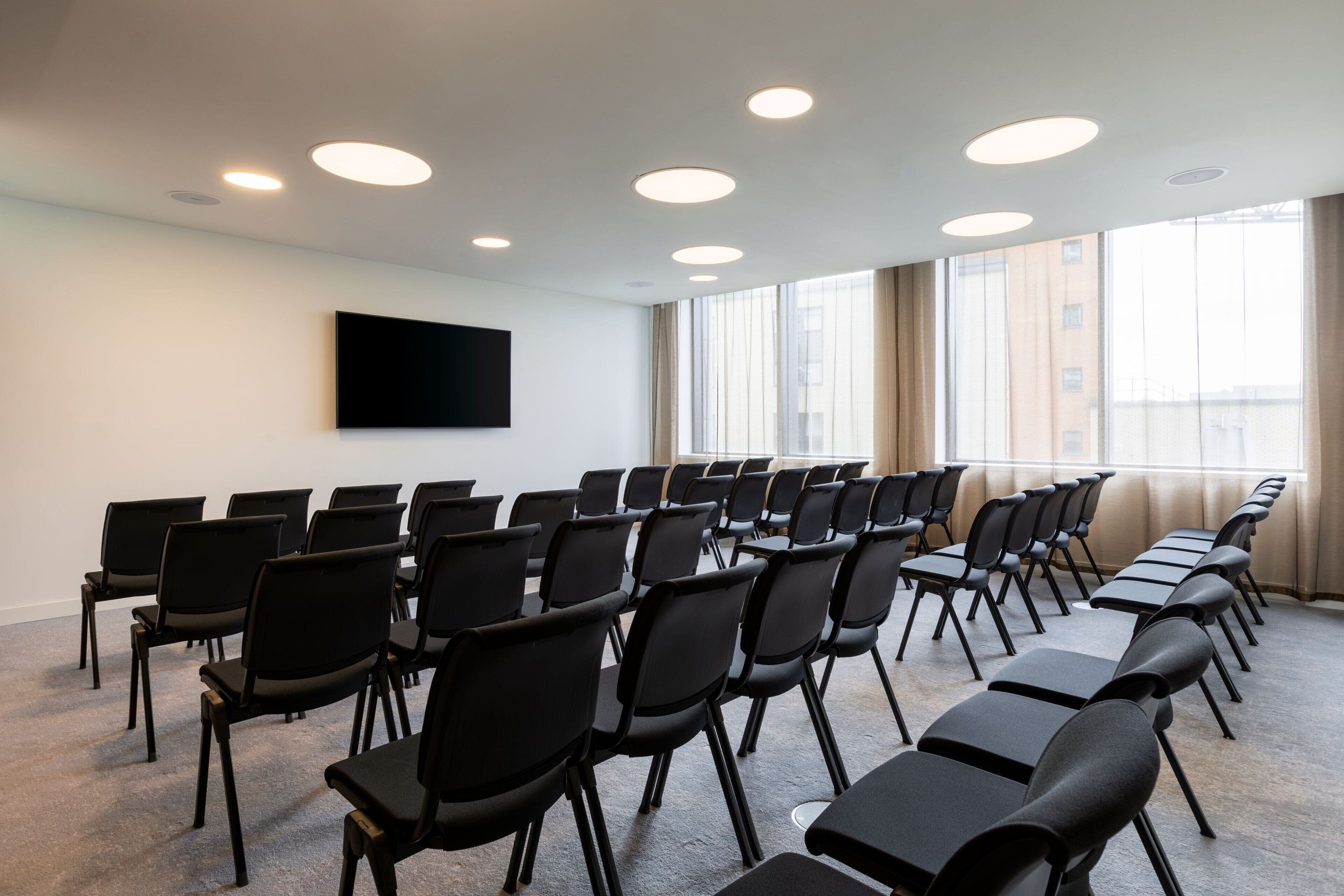 A conference room with rows of black chairs, a flat-screen TV on the wall, and large windows with curtains.