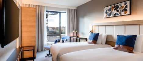 A hotel room with two beds, a desk, a chair, a flat-screen TV, and a large window with curtains.