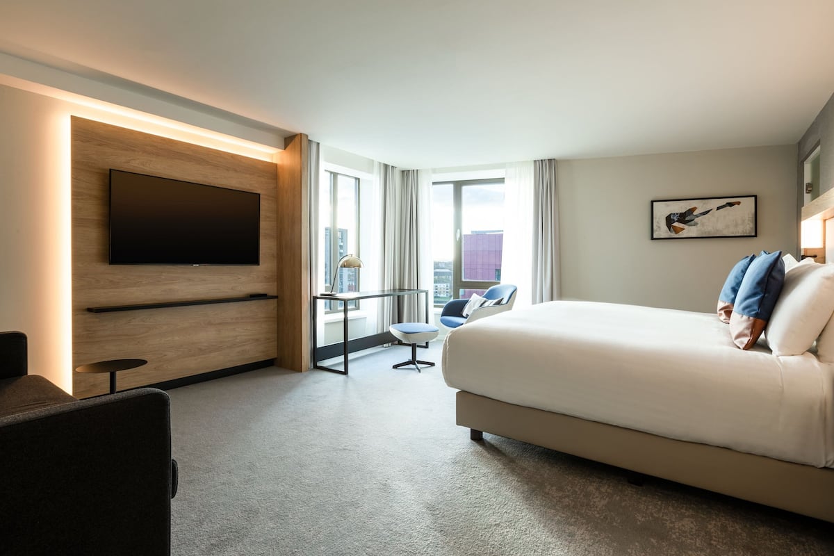 A modern hotel room with a large bed, a flat-screen TV mounted on a wooden panel, a small table, a chair, and a city view through large windows.
