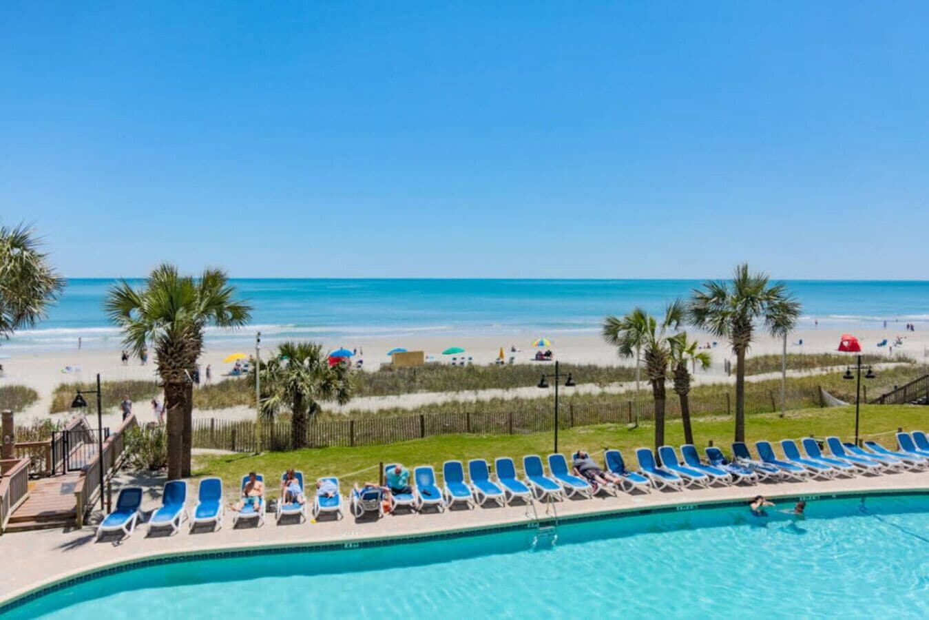 A poolside view with a beach in the background, lounge chairs, and palm trees.