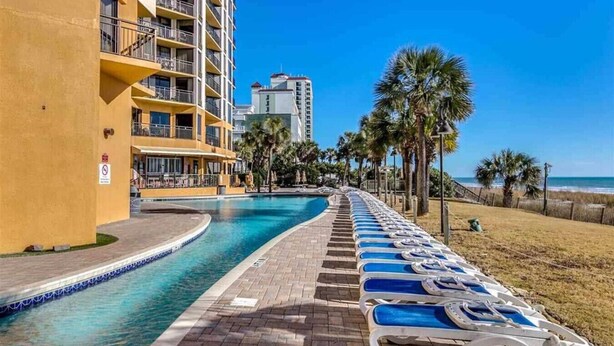 A hotel pool area with a clear view of the ocean, a palm tree-lined walkway, and a building with balconies. - 210Sea Gem. Oceanfront. Pet Friendly (Myrtle Beach)