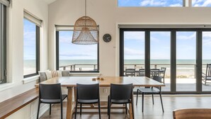 A modern dining area with a wooden table and chairs, large windows with a view of the beach, and a hanging light fixture. - New! Pool! Sleeps 14, Sol de Mar Beach, huge views, modern, duplex unit A (Carolina Beach)