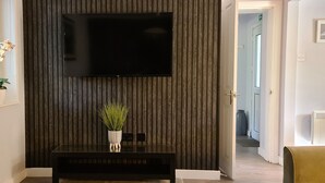 Een moderne kamer met een flatscreen-tv aan een gestructureerde muur, een glazen tafel met een potplant en een deur naar een andere ruimte.