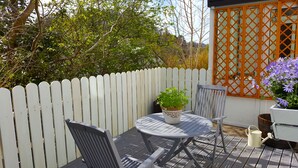 Een terras met een witte picket hek, een ronde tafel, twee stoelen, een bloempot en een houten vat.