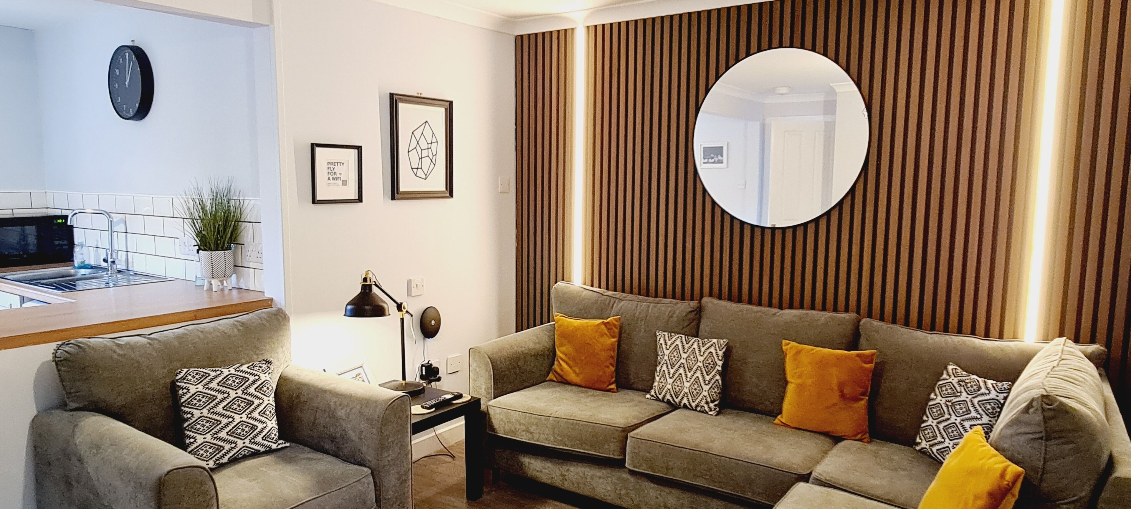 Una sala de estar moderna con un sofá, una mesa de centro, una mesita auxiliar, un espejo montado en la pared y una zona de cocina con microondas y una planta.