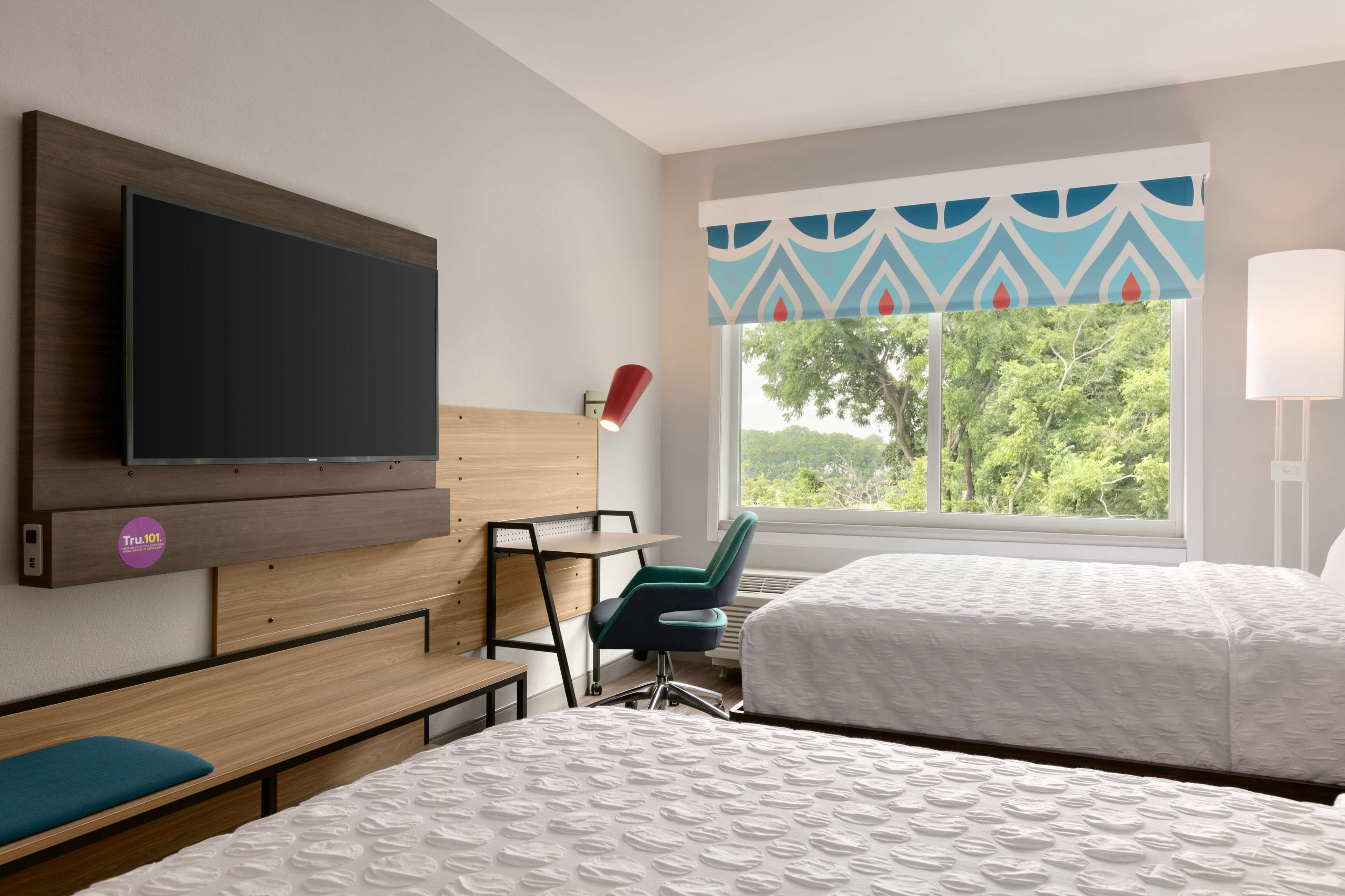 A hotel room with a bed, a desk, a chair, a TV, and a window with a view of trees.