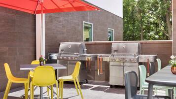 Un patio exterior con una parrilla, sillas amarillas y un paraguas rojo.