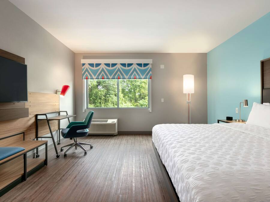 Une chambre d’hôtel comprenant un lit, un bureau avec une chaise, une télévision, une fenêtre donnant sur des arbres et une lampe.