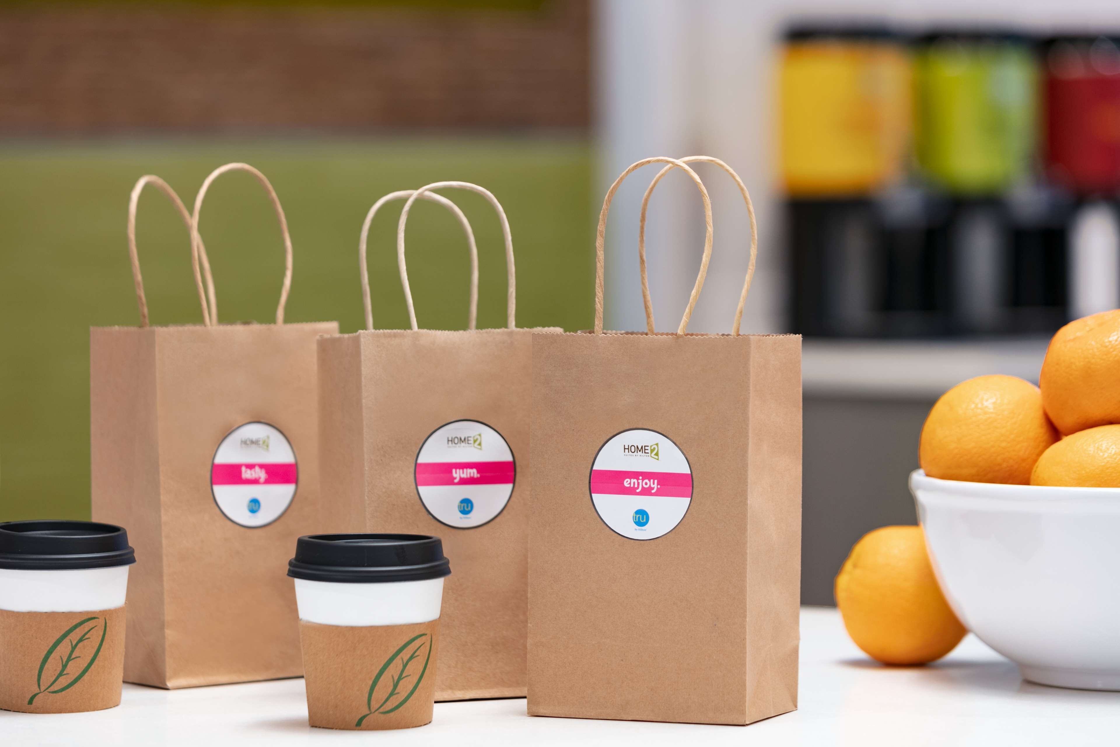 Three brown paper coffee bags with handles and lids, a bowl of oranges, and a blurred background with shelves.