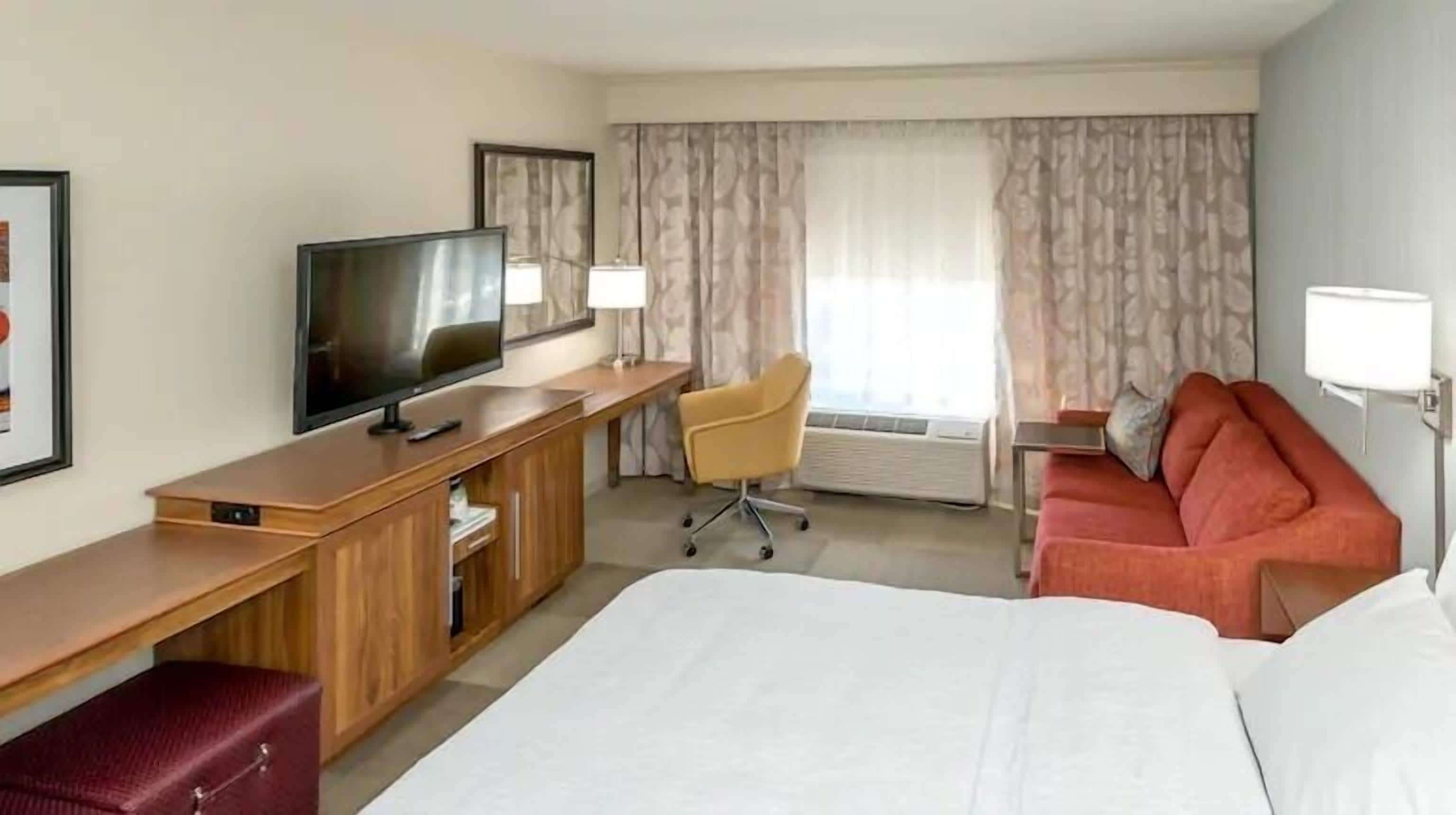 Habitación de hotel con una cama grande, un escritorio, un televisor de pantalla plana, una silla, un sofá y una ventana con cortinas.