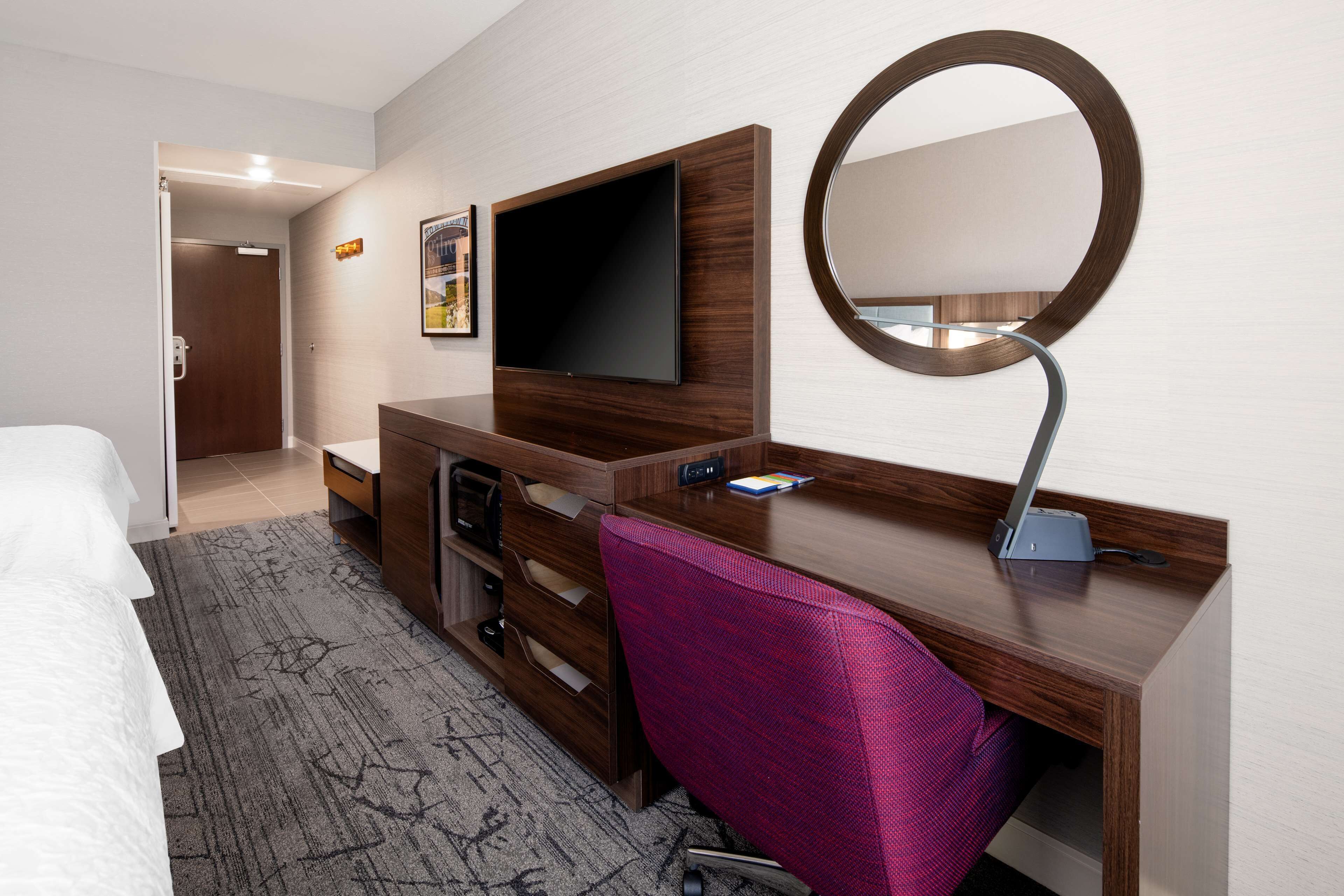 A hotel room with a bed, a desk with a flat-screen TV, a desk lamp, a chair, and a large mirror.