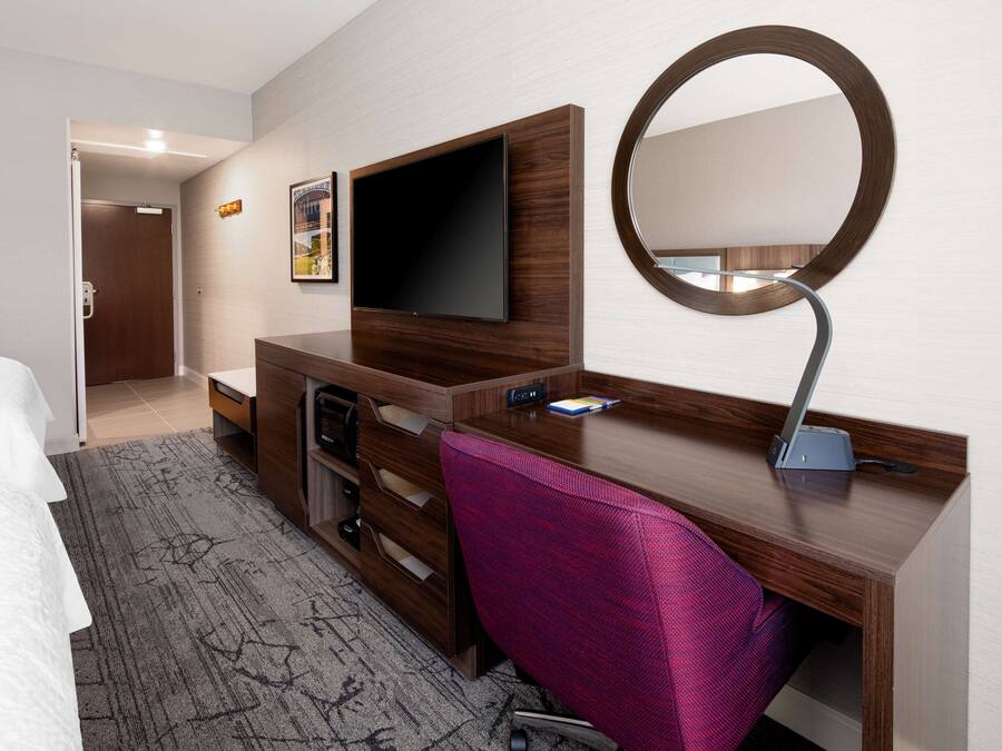 A hotel room with a bed, a desk with a flat-screen TV, a desk lamp, a chair, and a large mirror.