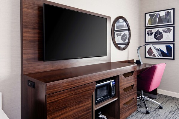 A modern room with a flat-screen TV mounted on a wooden cabinet, a microwave below it, and a pink office chair.