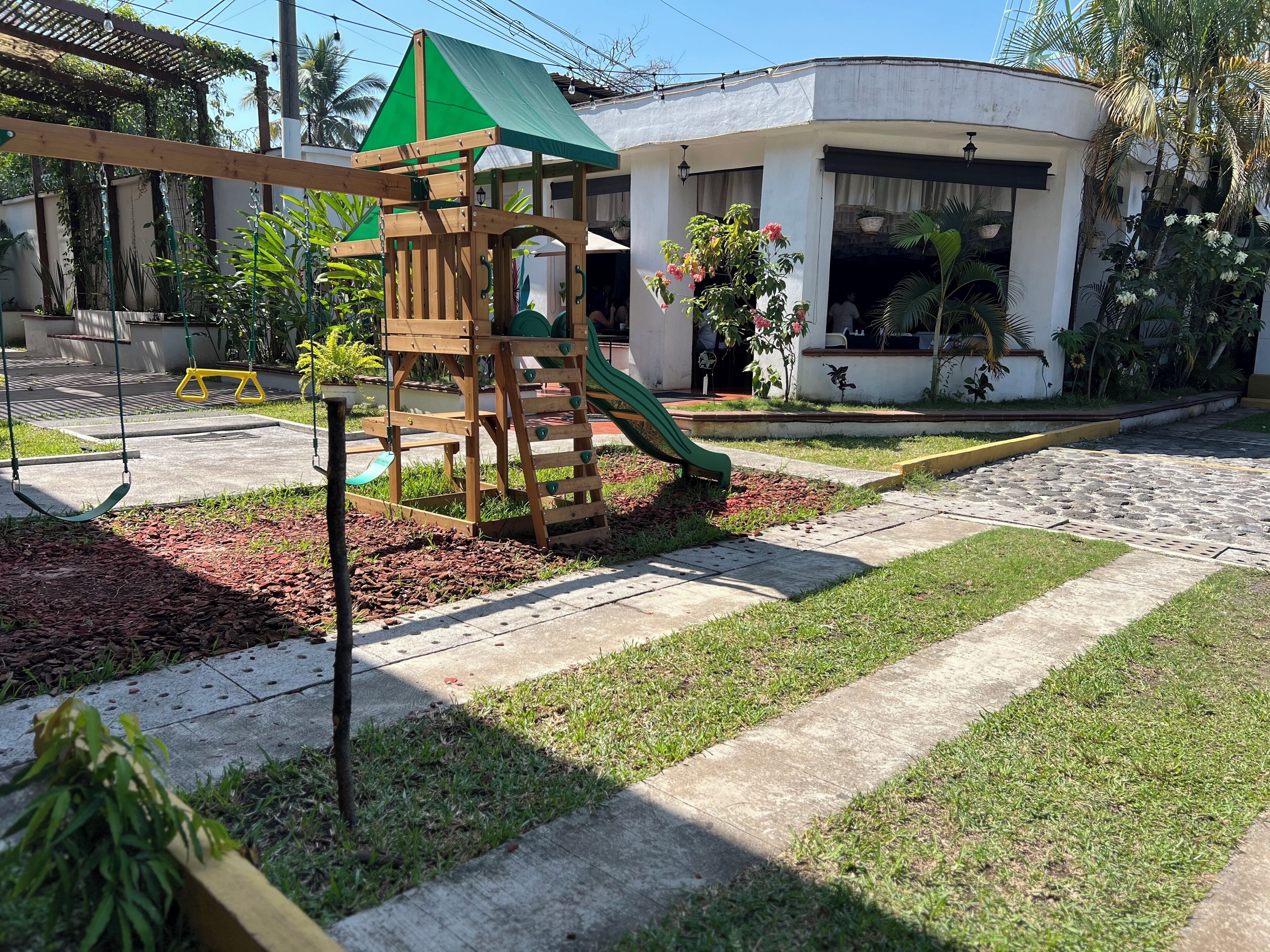 A playground with a slide and swings, surrounded by a garden with various plants and a building in the background.