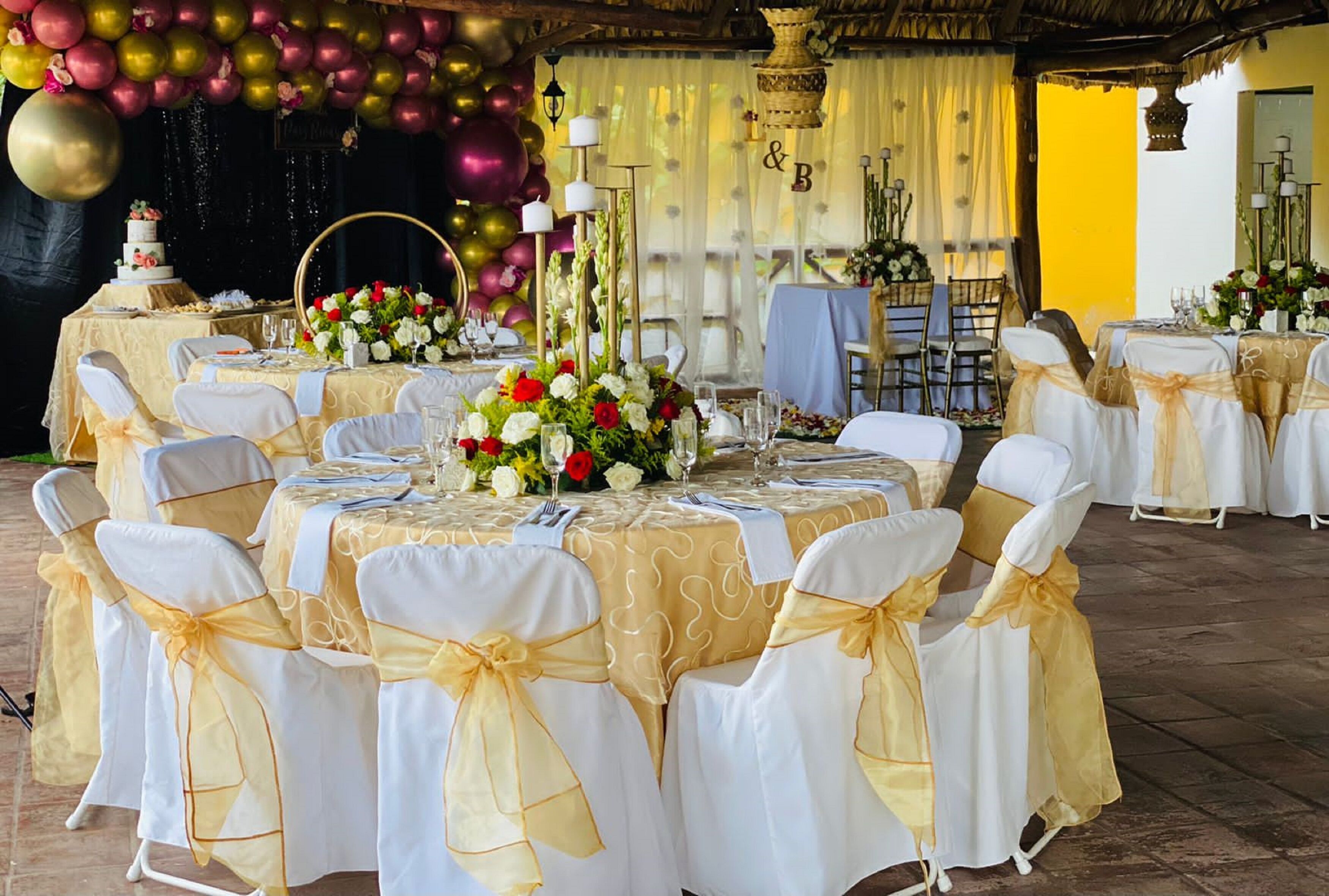 Un salón de banquetes con mesas redondas dispuestas para un evento formal, con manteles blancos y dorados, sillas con cintas doradas y centros de mesa florales.