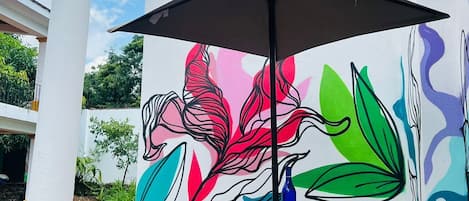 Um terraço ao ar livre com mesa e cadeiras, um guarda-sol preto e um mural colorido na parede.