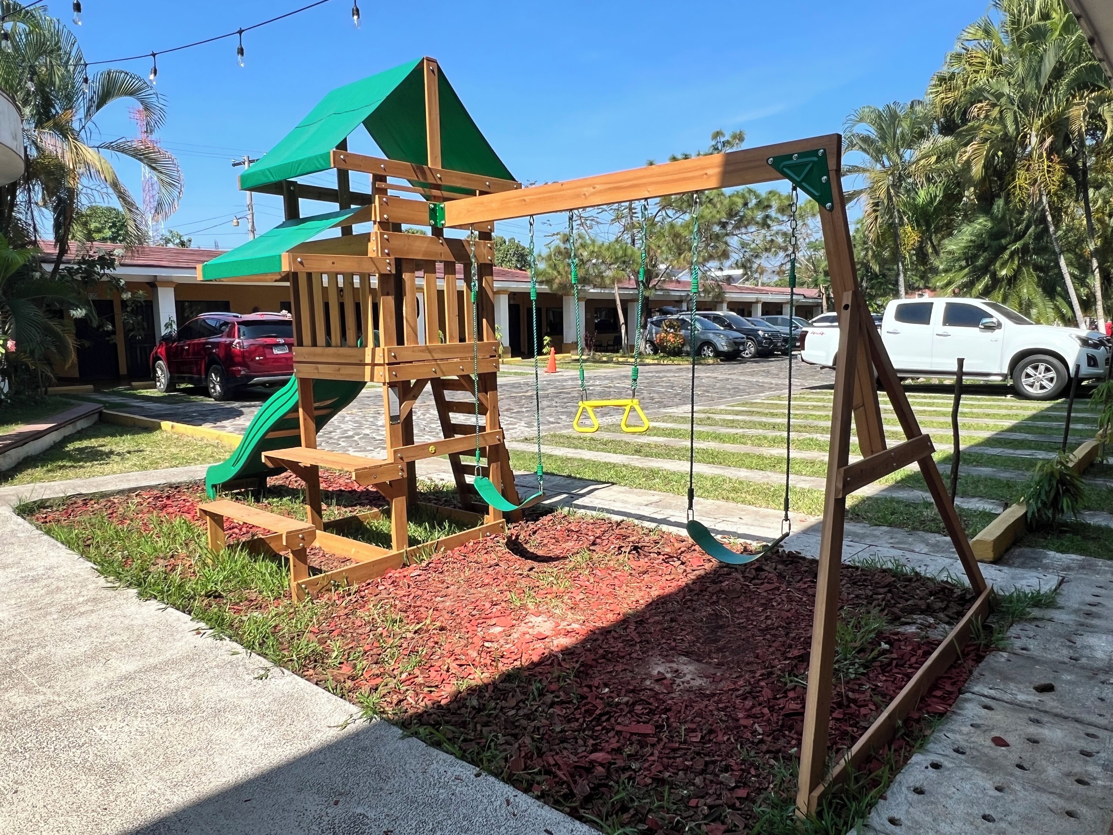 A playground with a wooden play structure, including a slide and swings, surrounded by a landscaped area with mulch and grass.