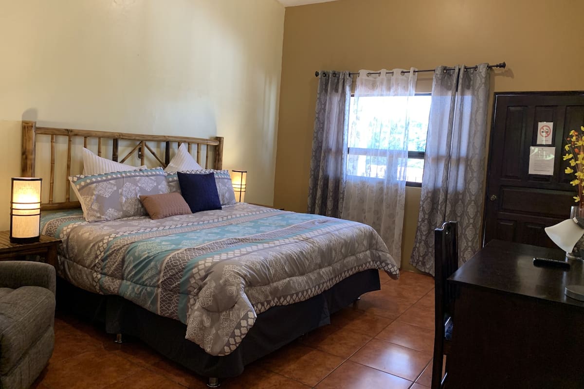 A bedroom with a wooden headboard, a patterned bedspread, two bedside lamps, and a window with curtains.