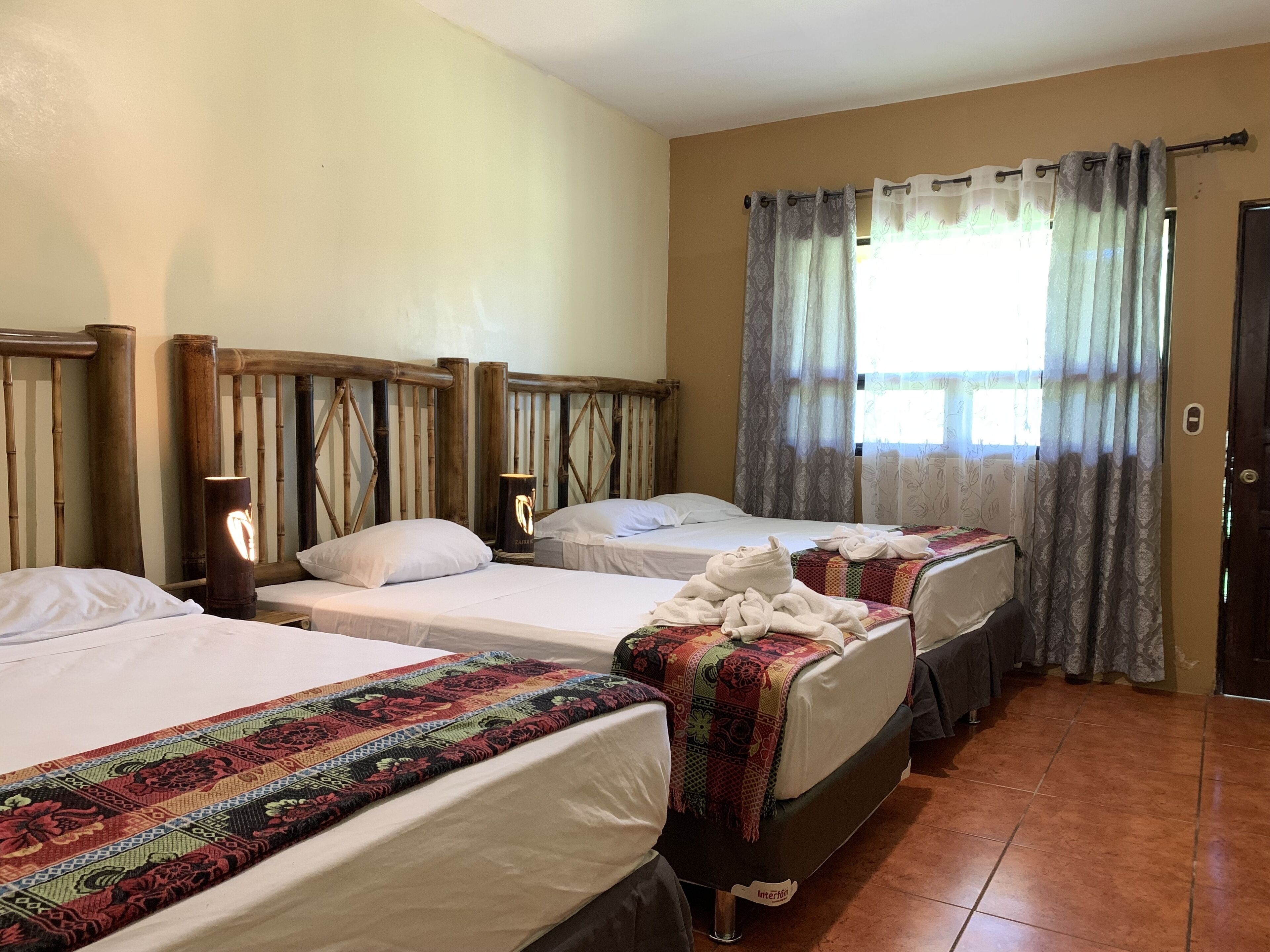 A hotel room with two single beds, a wooden headboard, a bedside table with a lamp, and a window with patterned curtains.