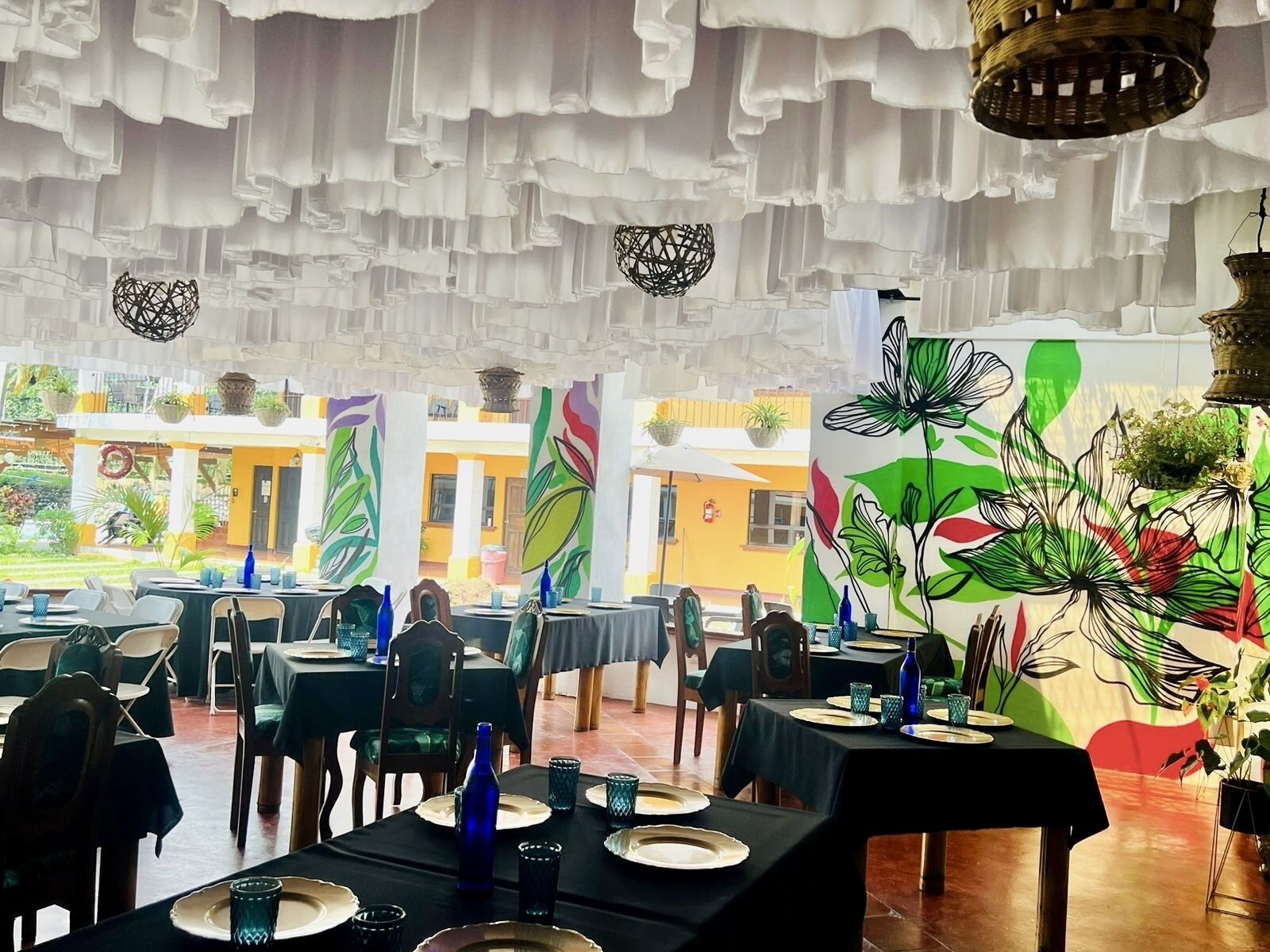 Een restaurant met gedekte tafels, versierd met hangende decoraties en een muurschildering met bloemmotieven.