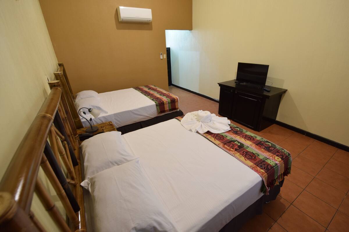 A hotel room with two single beds, a small desk with a television, and a wall-mounted air conditioner.
