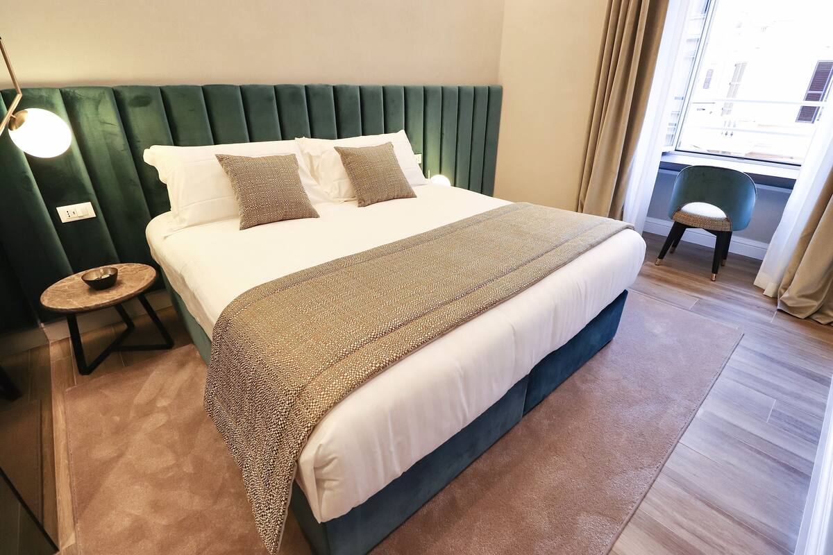 A neatly made bed with a green upholstered headboard, a bedside table with a lamp, and a view of the outside through a window.