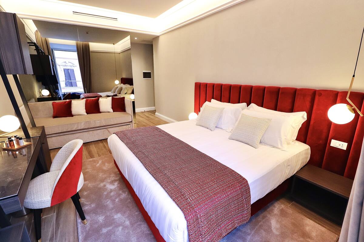 A hotel room with a large bed, a desk with a chair, a mirror, and a red accent wall.