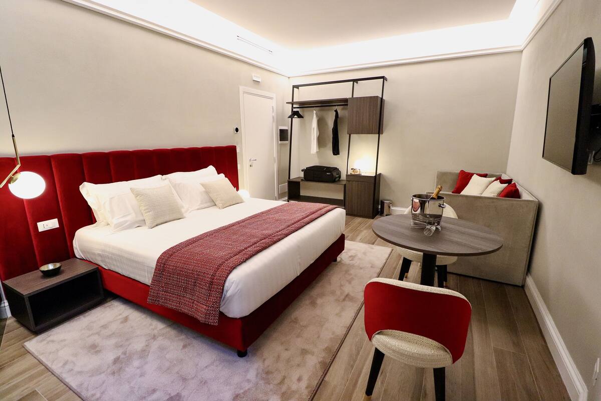 A modern hotel room with a red upholstered bed, a small table with a bottle, a red chair, and a sofa with red cushions.