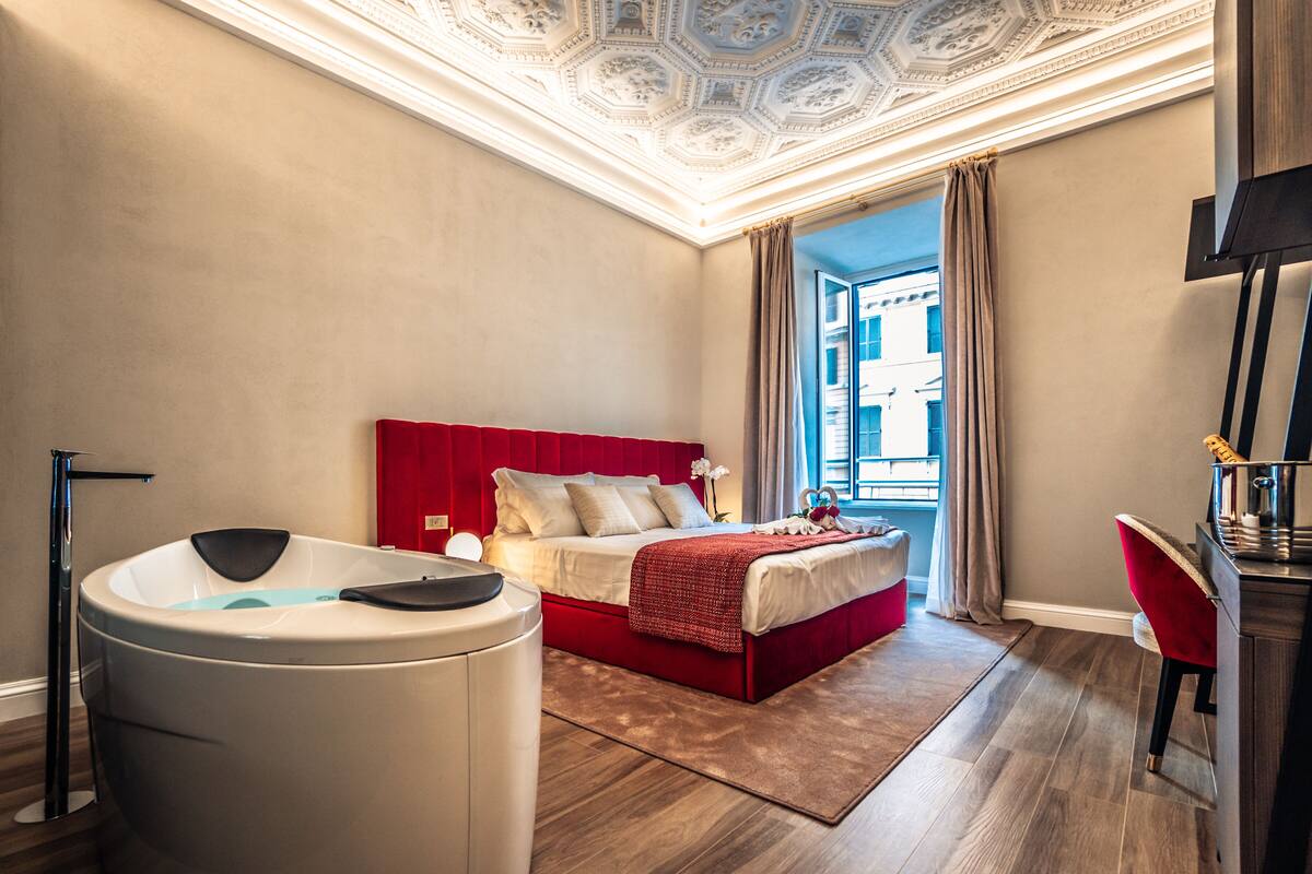 A modern hotel room with a red bed, a bathtub, and a view of a building through a window.