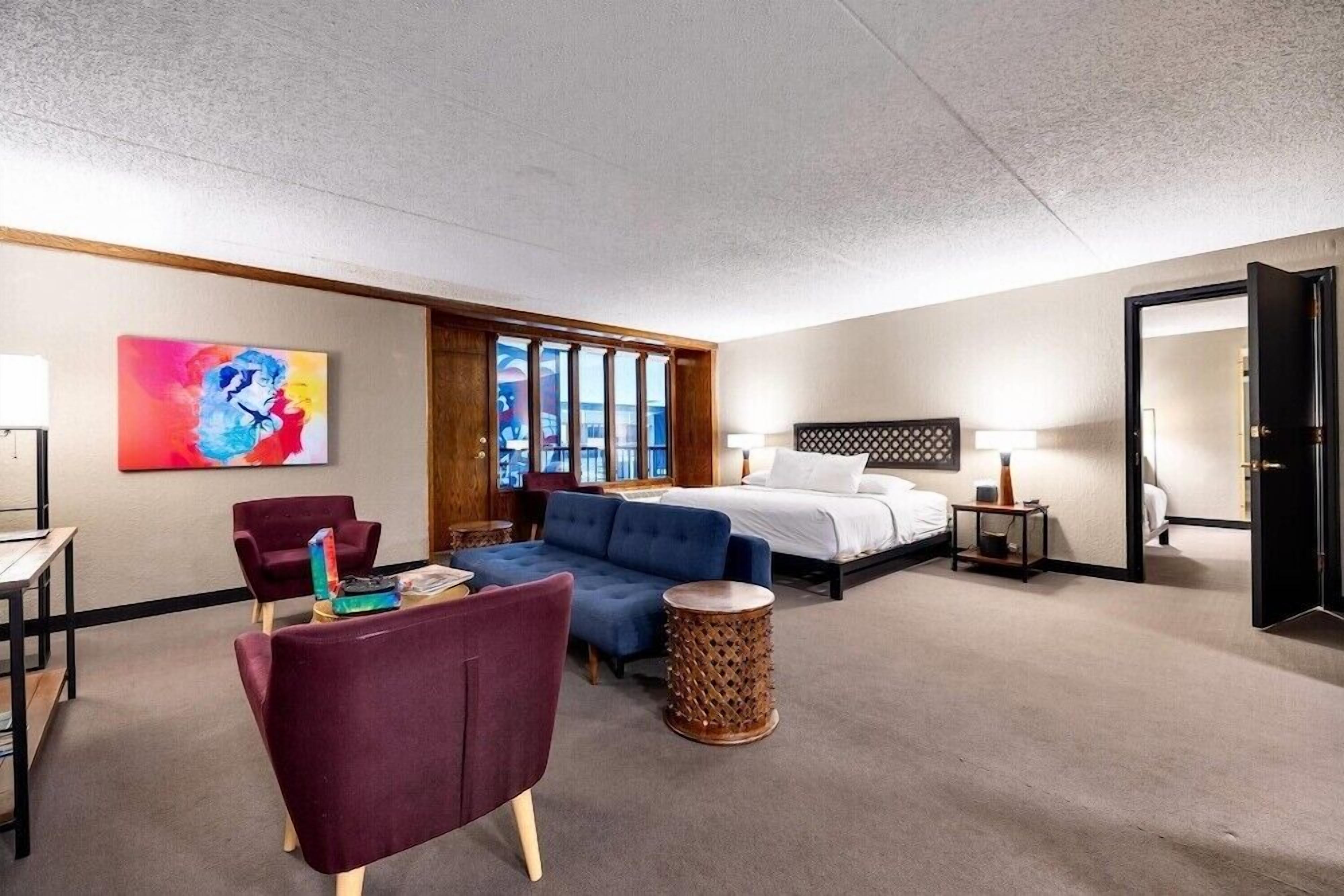 A hotel room with a bed, a sofa, a chair, a small table, and a painting on the wall.