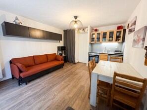Een knus woongedeelte met een bank, eettafel, stoelen, een kitchenette en een plafondlamp.