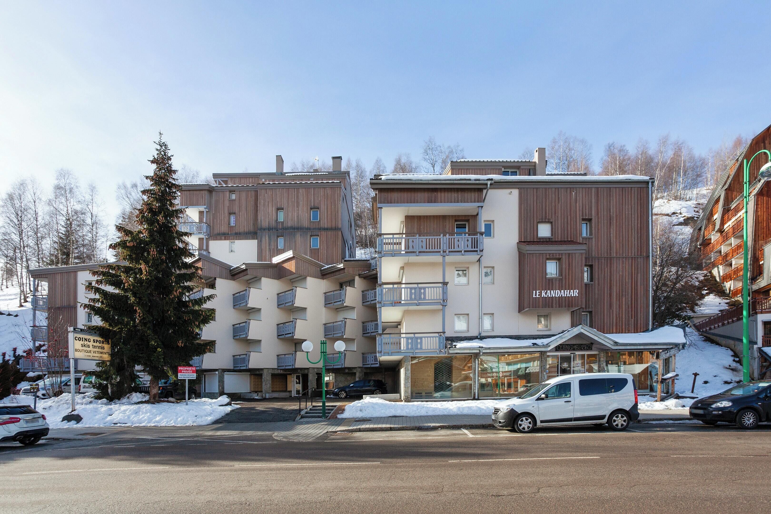 Un hôtel à plusieurs étages doté de balcons, un panneau indiquant la présence d’une station de ski, et un espace de stationnement enneigé.